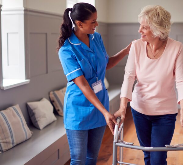 Senior Woman At Home Using Walking Frame Being Helped By Female Care Worker In Uniform